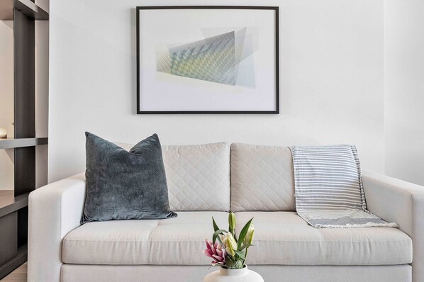 A living room with a light-colored sofa, a framed abstract painting, and a vase with flowers.