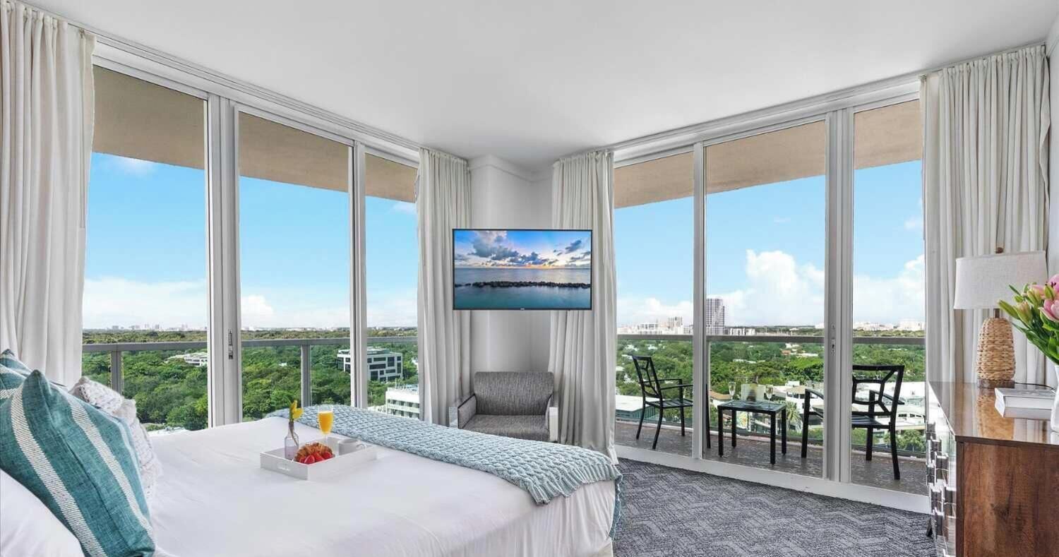A hotel room with a large bed, a TV, a balcony with chairs, and a city view.