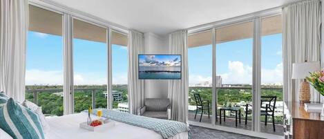 A hotel room with a large bed, a TV, a balcony with chairs, and a city view.