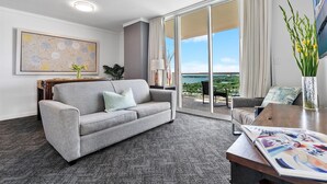Una habitación de hotel moderna con un sofá gris, una mesa de centro, un amplio ventanal con vistas al exterior y un cuadro en la pared.