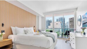 A modern hotel room with a large bed, a wooden slat wall, a desk, and a view of high-rise buildings. - Modern Residence with Unforgettable Grove Views (Miami)