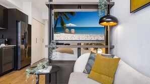 A modern living room with a large flat-screen TV displaying a beach scene, a sofa with a mustard-colored pillow, a small table with a plant, and a sleek kitchen with dark cabinetry.