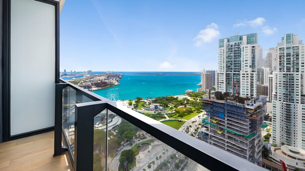 A high-rise building with a glass balcony overlooking a coastal cityscape, including a large body of water and a Ferris wheel.