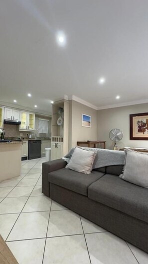 A living room with a sofa, a fan, and a kitchen in the background. - Diani Doric Cottages (Diani Beach)