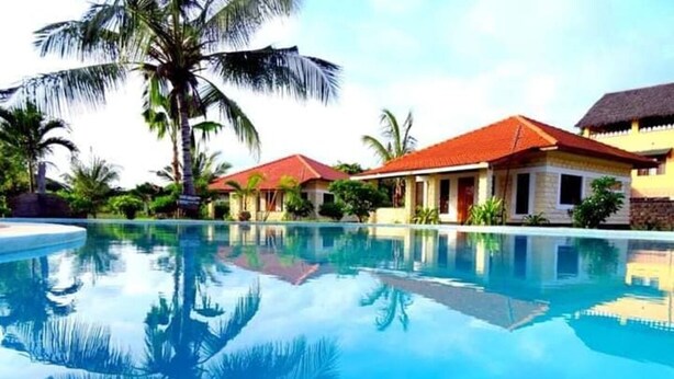 A swimming pool with a reflection of palm trees and a building with a red roof. - Diani Doric Cottages (Diani Beach)