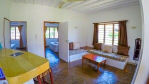 A spacious living area with a sofa set, a wooden table, and a ceiling fan. - Diani Doric Cottages (Diani Beach)