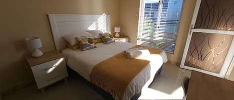 A bedroom with a large bed, bedside tables, a window with blinds, and artwork on the wall.