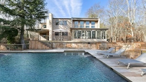 A modern house with a swimming pool, a deck with sun loungers, and a large window overlooking the pool.