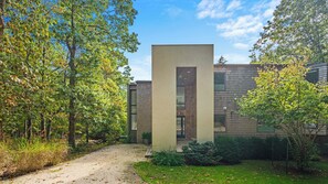 A modern building with a central vertical structure, surrounded by trees and a gravel path.