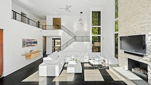 A spacious living area with a white sofa set, a coffee table, and a fireplace. A staircase leads to an upper level, and a TV is mounted on the wall.