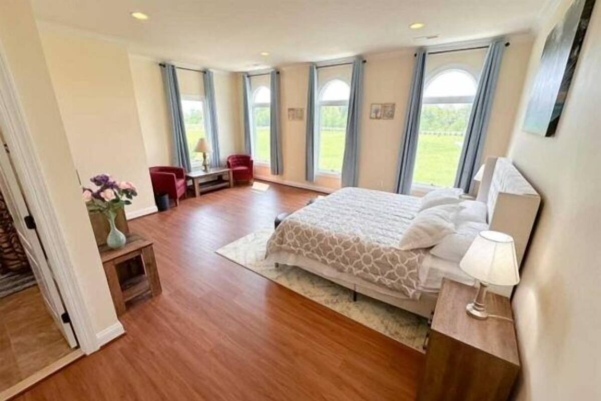 A spacious bedroom with a large bed, a desk, and a chair. There are multiple windows providing natural light and a view of the outdoors.