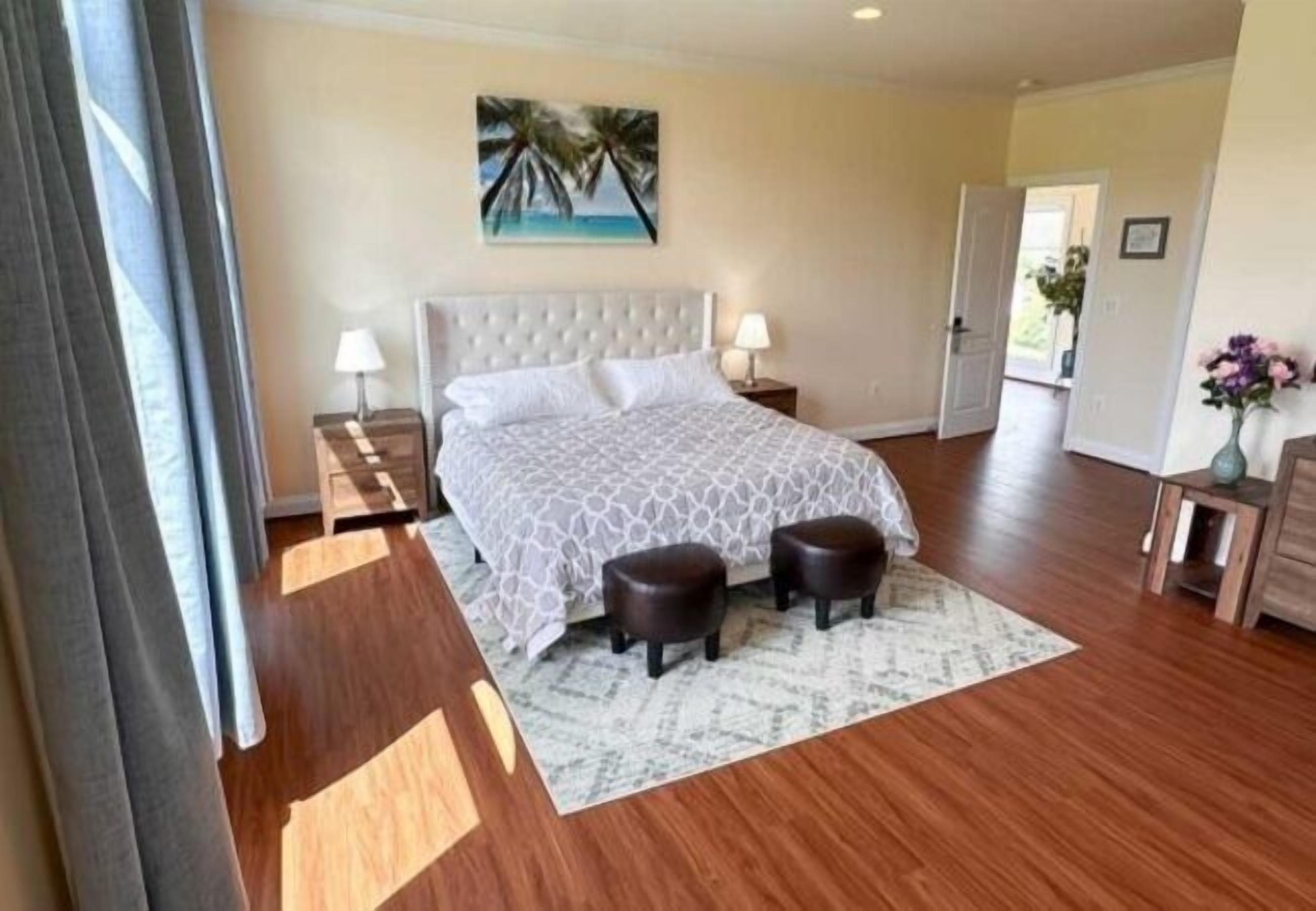 A hotel room with a large bed, two bedside tables, a nightstand, a painting of palm trees, and a wooden floor.