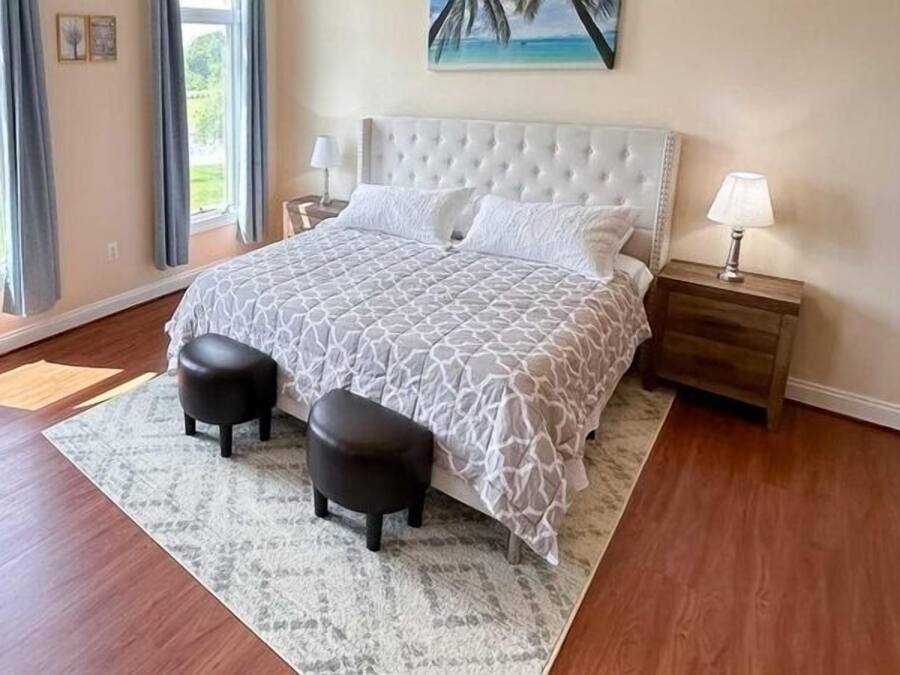 Quarto com uma cama grande, duas mesas de cabeceira, uma luminária, janela com cortinas e vista para o verde lá fora.