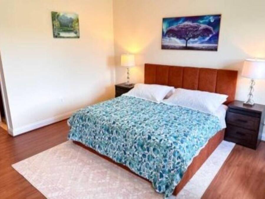 Un dormitorio con una cama, dos mesitas de noche, una mesita auxiliar, un cuadro colgado en la pared y una puerta.