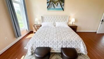 Un dormitorio con una cama grande, dos mesitas de noche, un cuadro de palmeras y vistas al exterior.