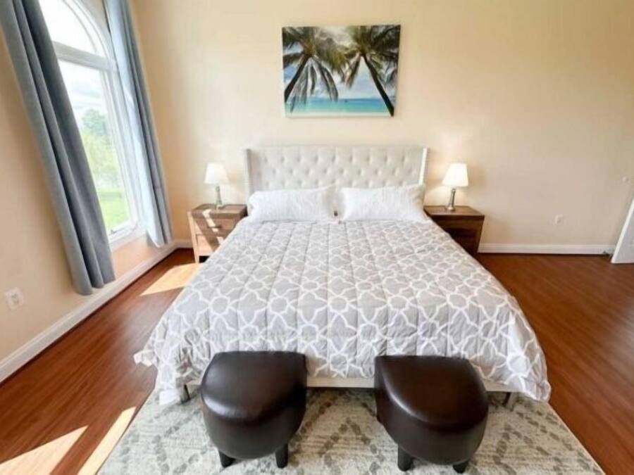 Un dormitorio con una cama grande, dos mesitas de noche, un cuadro de palmeras y vistas al exterior.