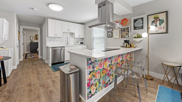 A modern kitchen with white cabinets, a stainless steel range hood, and a colorful backsplash. There are bar stools and a small round table with a lamp. - Optimist Abode 1: Chic Stay on Greenway, less than 7 min to NoDa-Midwood-Uptown (Charlotte)