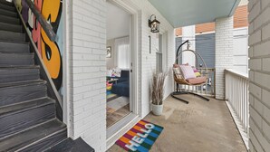A covered porch with a white brick wall, a black staircase, a colorful 'HELLO' mat, a hanging chair, and a potted plant. - Optimist Abode 1: Chic Stay on Greenway, less than 7 min to NoDa-Midwood-Uptown (Charlotte)
