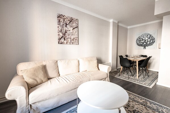 A living room with a beige sofa, a round white coffee table, and a dining area with a table and chairs.