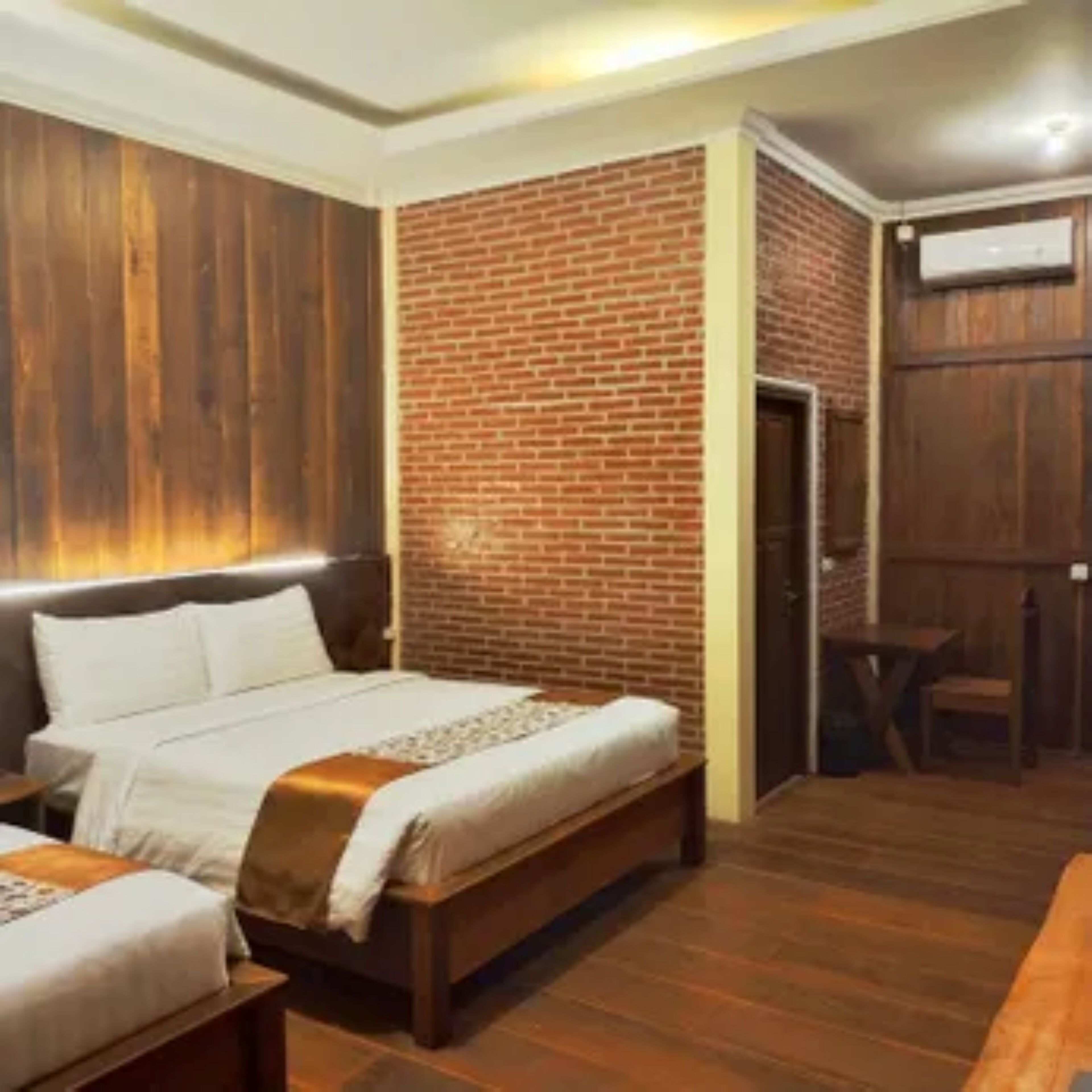 A hotel room with a wooden floor, a brick accent wall, a bed with a headboard, and a small table with a chair.