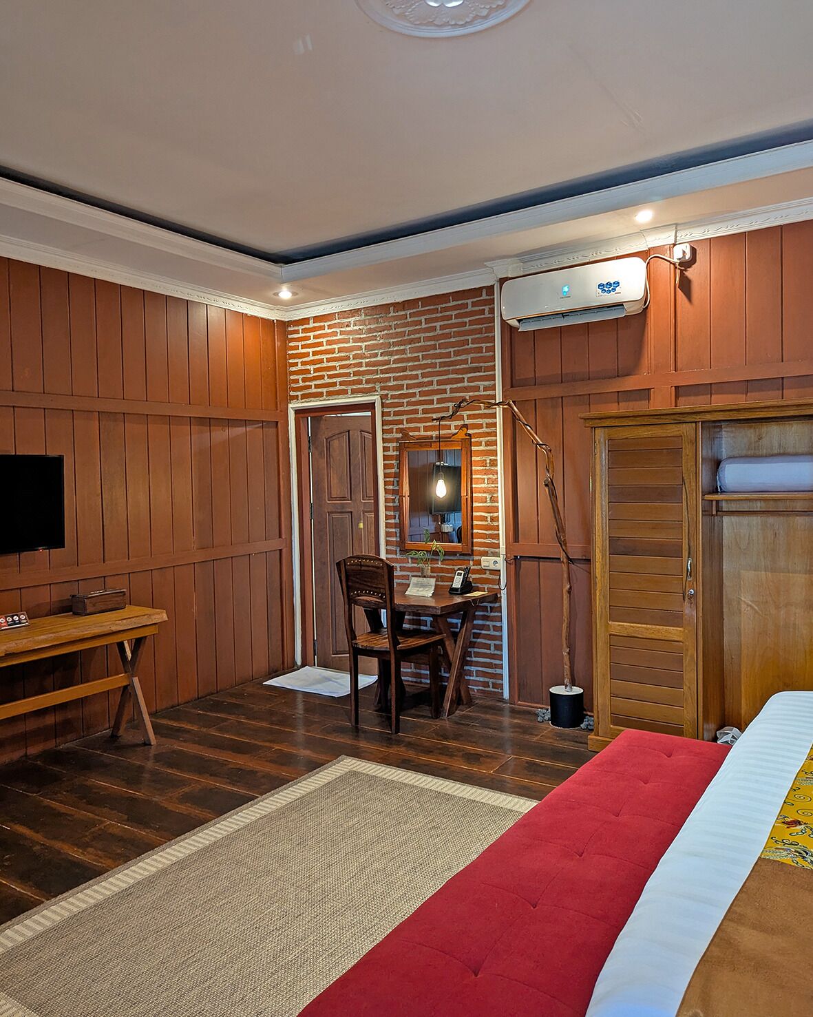 Una habitación con un revestimiento de madera, pared de ladrillo decorativa, un escritorio con una silla, cama con detalles rojos y un televisor a la izquierda.