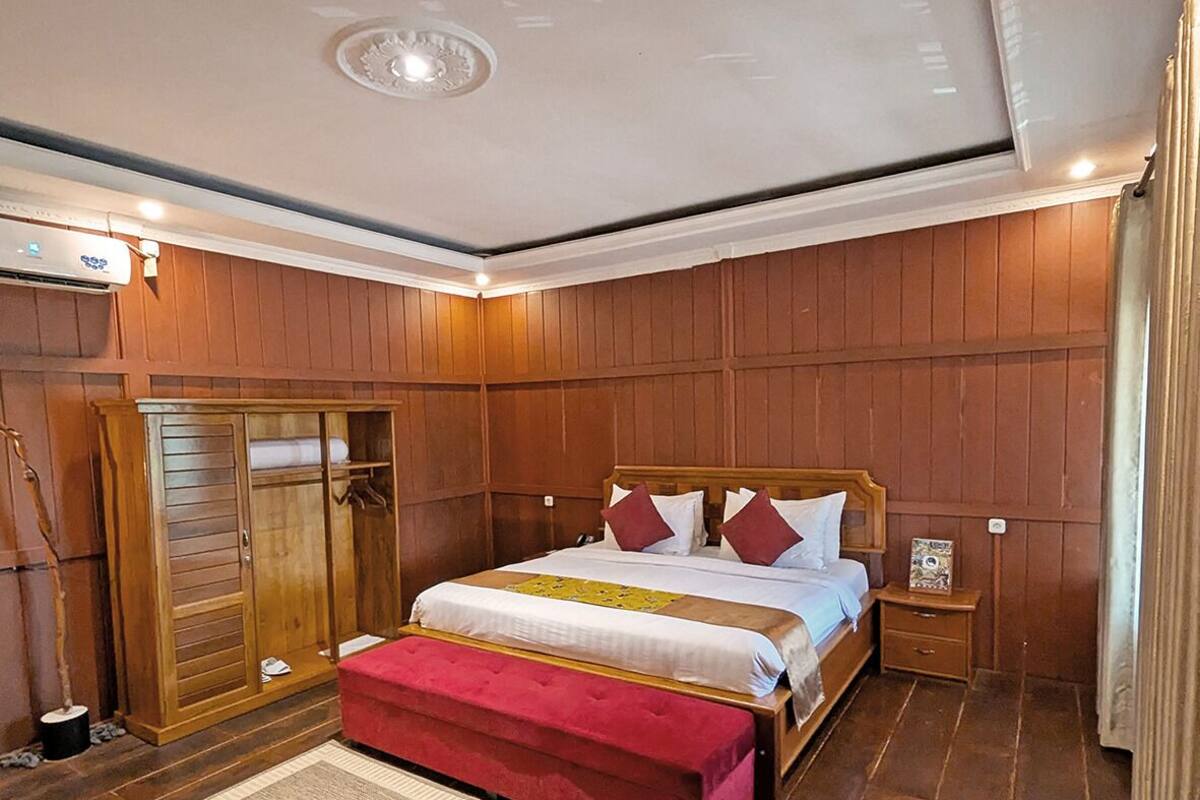 A bedroom with a wooden panel wall, a bed with a red upholstered bench, and a small bedside table.