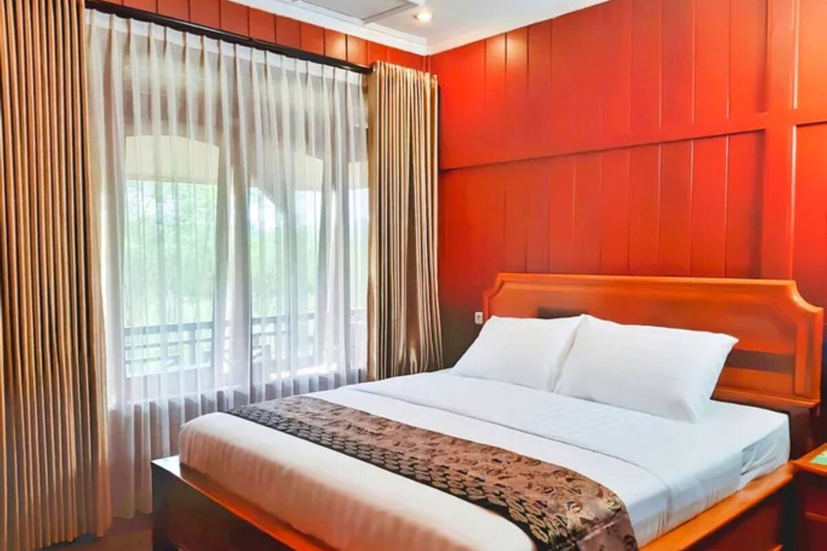 A hotel room with a wooden bed, white bedding, and a view of greenery through sheer curtains.