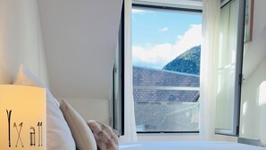 A bedroom with a bed, a nightstand with a lamp, and a window with a view of a building and hills. - Idéal Cure thermale à Cauterets - Near thermal baths & town center, 32 m² T2 (Cauterets)