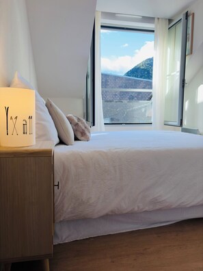 A bedroom with a bed, a nightstand with a lamp, and a window with a view of a building and hills.