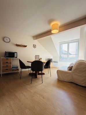 Quarto estilo loft com área de jantar, sofá, televisão, relógio e janela com vista.