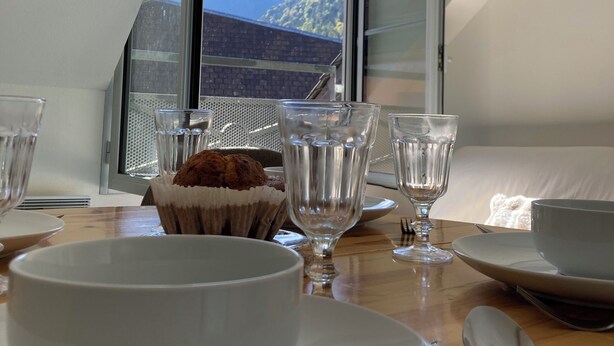 A dining table with a bowl, glasses, and a muffin in a paper cup, set against a mountain view through a window. - Idéal Cure thermale à Cauterets - Near thermal baths & town center, 32 m² T2 (Cauterets)