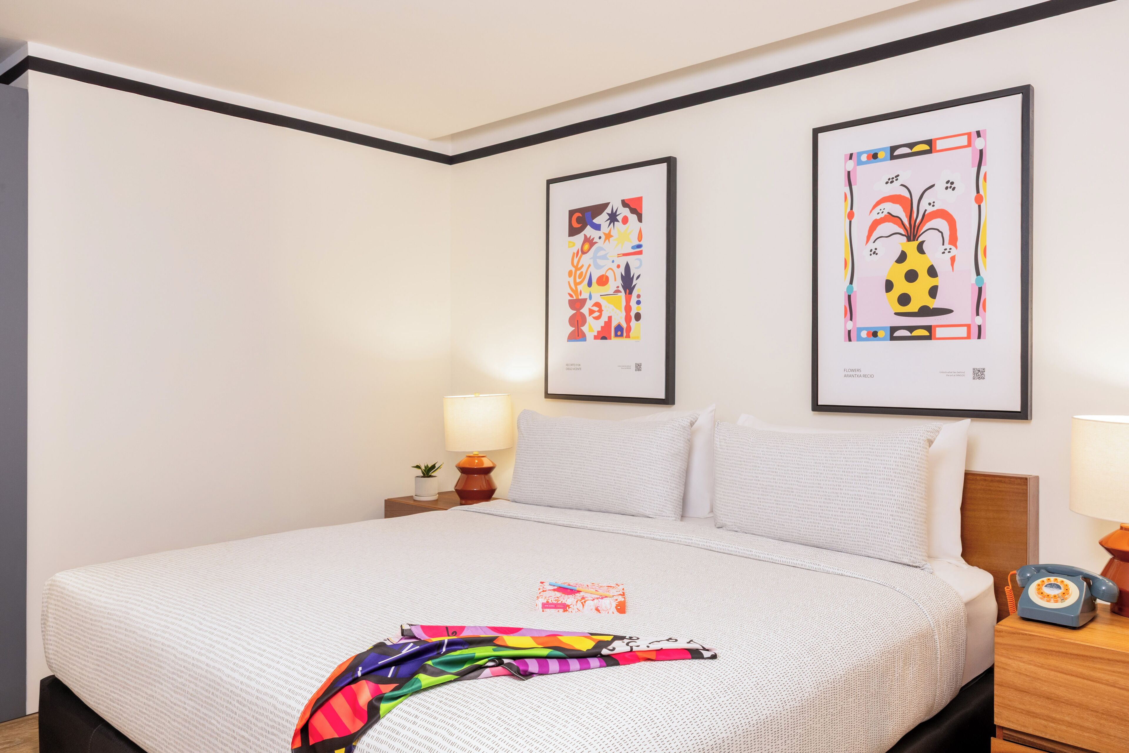 A hotel room with a large bed, two framed artworks on the wall, a bedside table with a lamp, and a telephone.