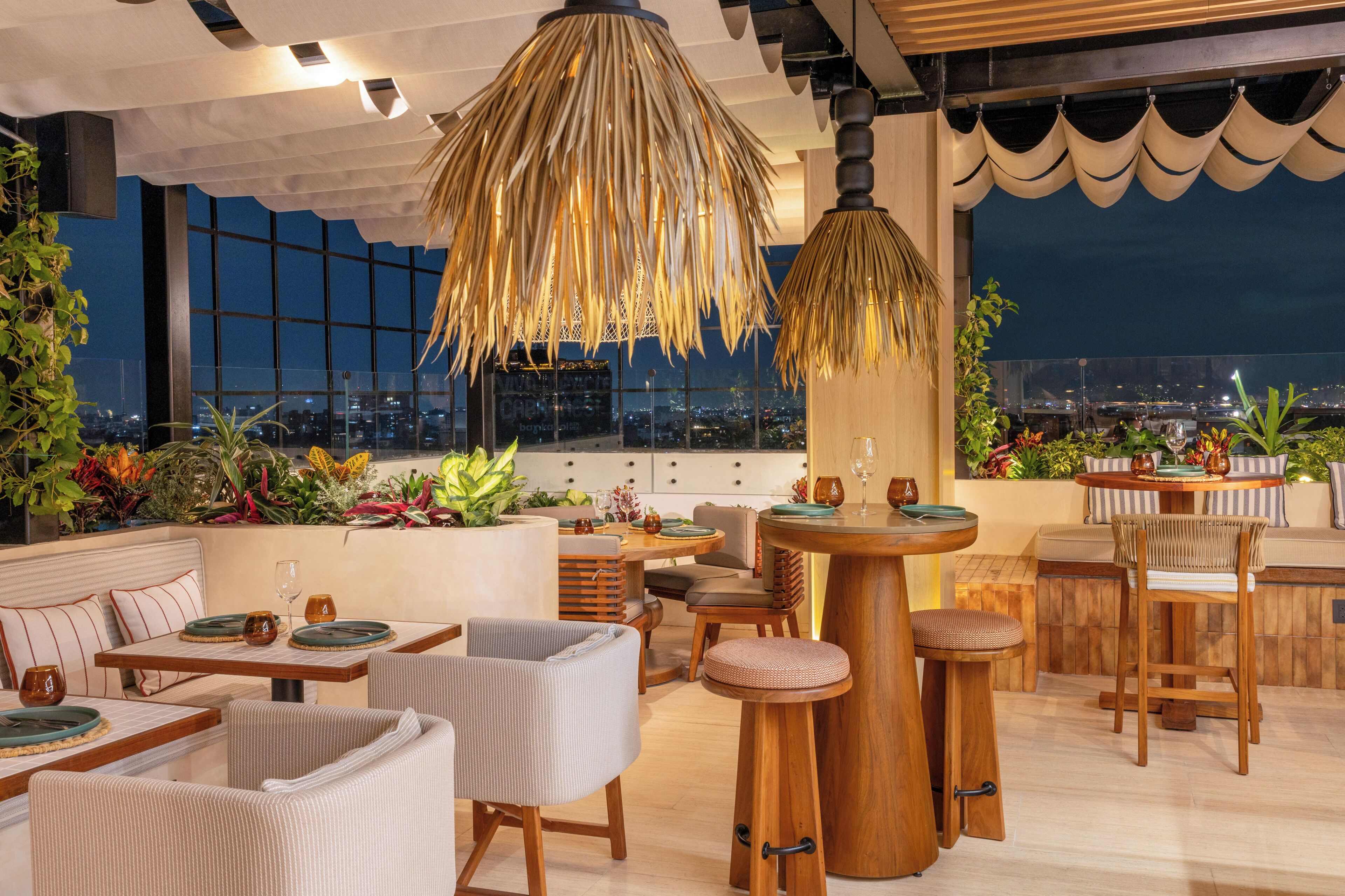 A rooftop bar with a view of the city at night, featuring a large hanging light fixture, seating arrangements, and a variety of plants.