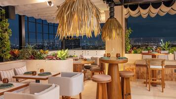 A rooftop bar with a view of the city at night, featuring a large hanging light fixture, seating arrangements, and a variety of plants.