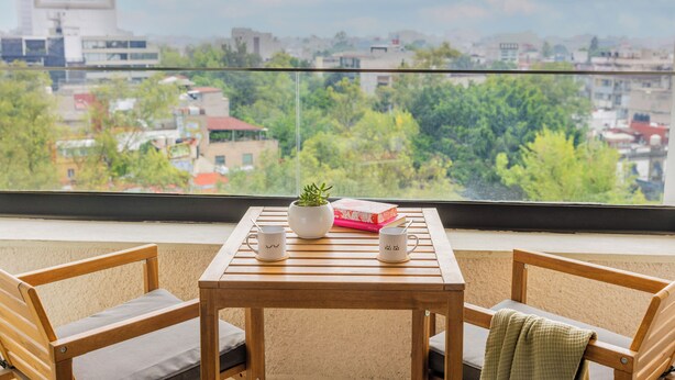 Una habitación con vistas a un paisaje urbano, una mesa con dos sillas, una planta en maceta y un libro.