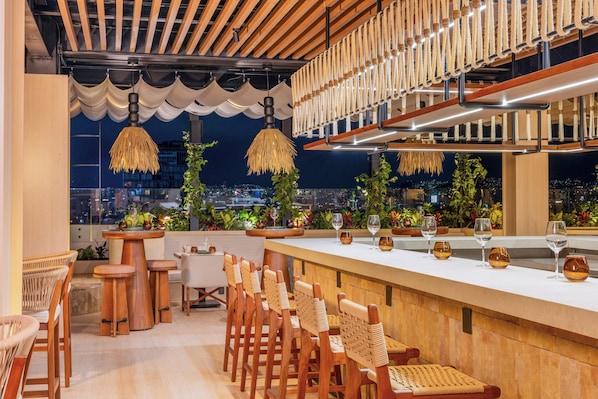 Un bar en la azotea con mobiliario de madera, luces colgantes y vistas a la ciudad.