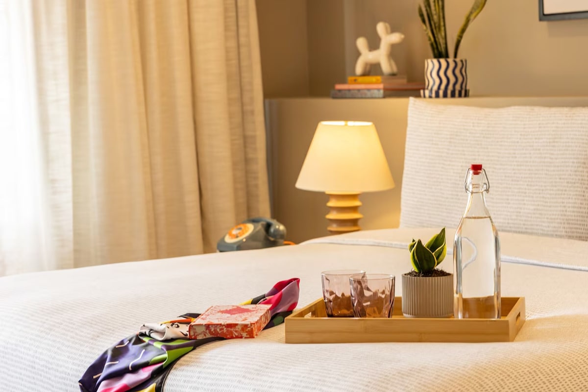 A neatly made bed with a tray holding a bottle, glasses, and a small plant.