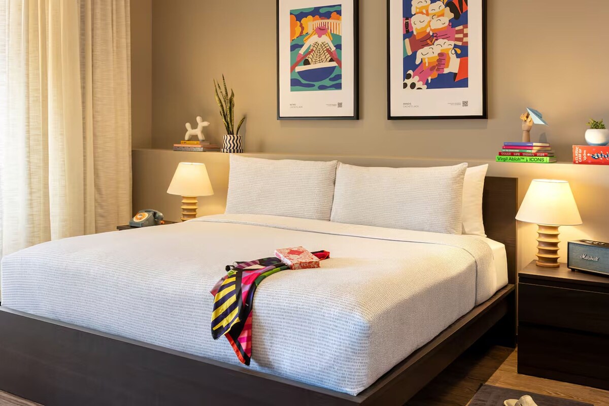 A hotel room with a large bed, two bedside lamps, a nightstand with a lamp, a small table with books, and two framed artworks on the wall.