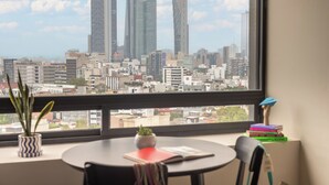 Una habitación con un gran ventanal que da a un paisaje urbano, una mesa redonda, una silla, una planta pequeña y una pila de libros.