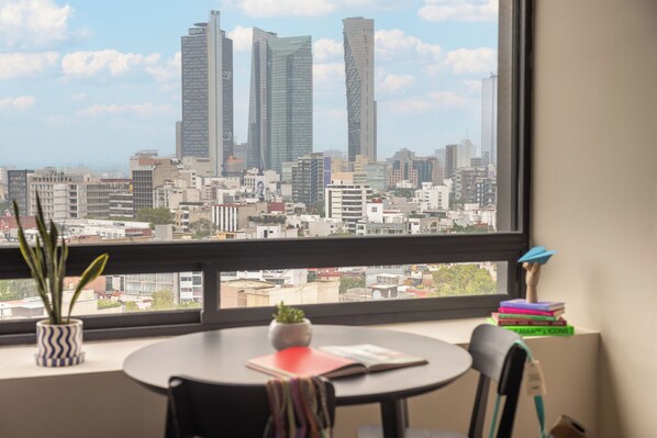 A room with a large window overlooking a cityscape, a round table, a chair, a small plant, and a stack of books.