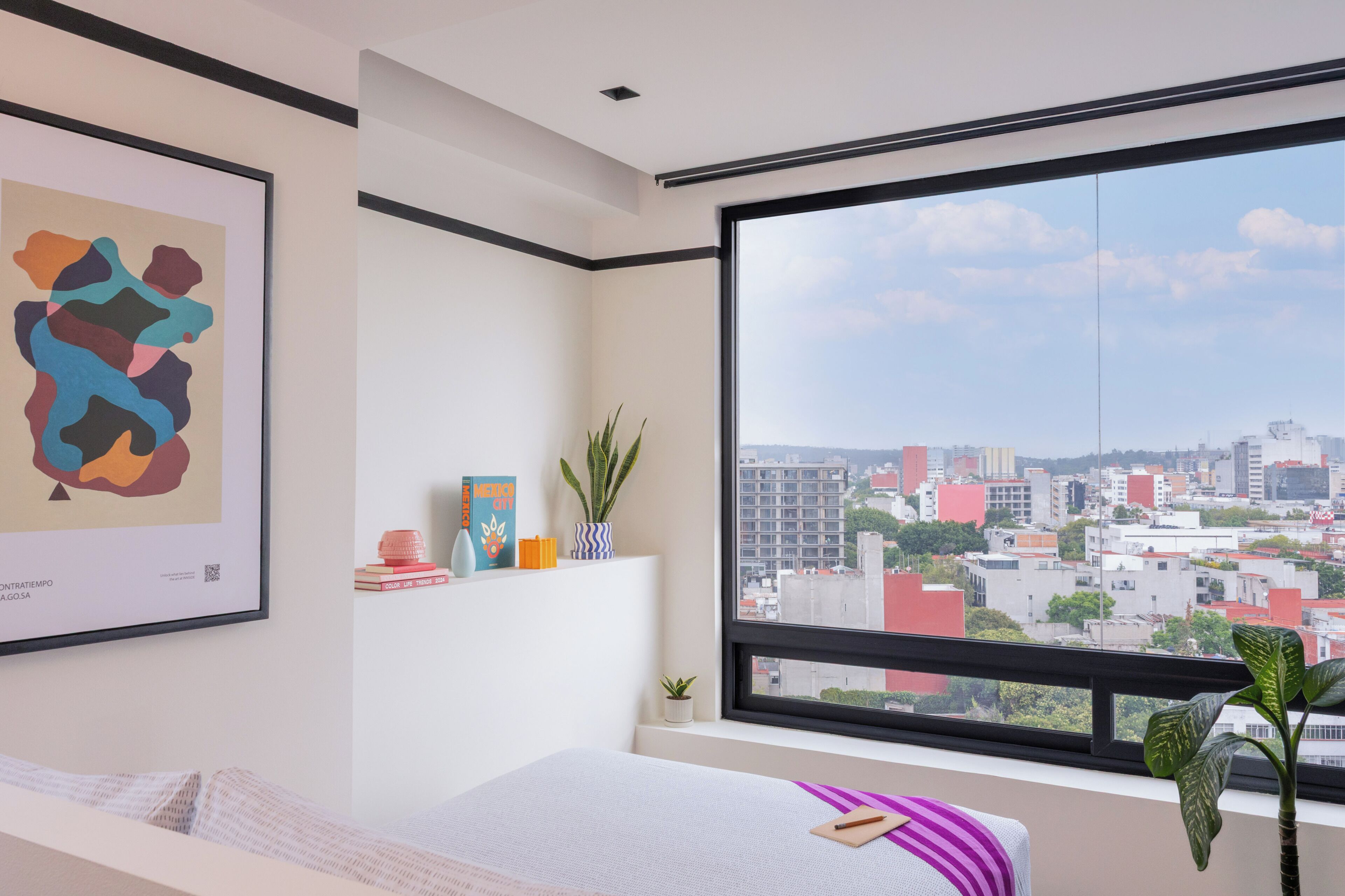 Una habitación moderna con un ventanal grande, una cama, un cuadro abstracto enmarcado y una planta pequeña.