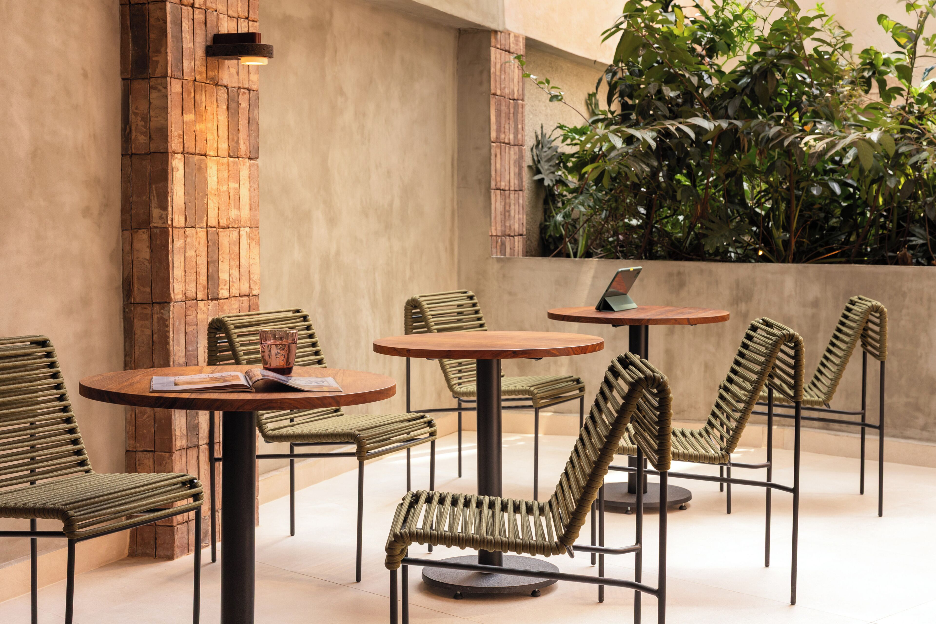 An outdoor seating area with round tables and woven chairs, surrounded by a stone wall and greenery.