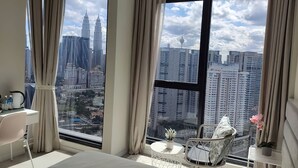 A modern hotel room with a large window offering a city view, a bed, a desk with a chair, and a small table with a plant.