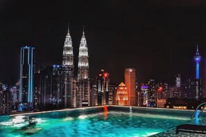 Nattebillede af en by med oplyste skyskrabere, heriblandt Petronas Towers, og en pool der spejler byens lys.