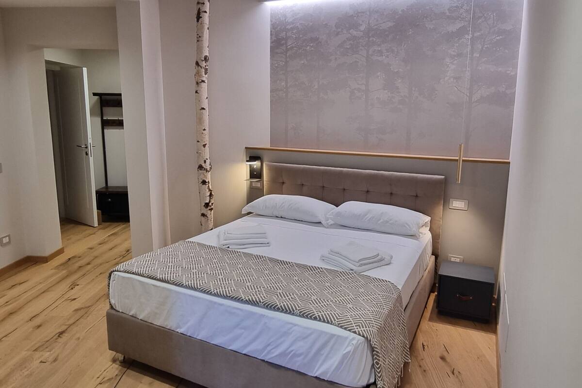A modern bedroom with a large bed, wooden flooring, a headboard with built-in lighting, and a view of trees through a window.
