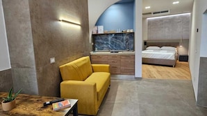 A modern bedroom with a bed, a yellow sofa, a small table with a plant, and a bathroom with a blue accent wall. - Il Piccolo Paradiso (Civitavecchia)