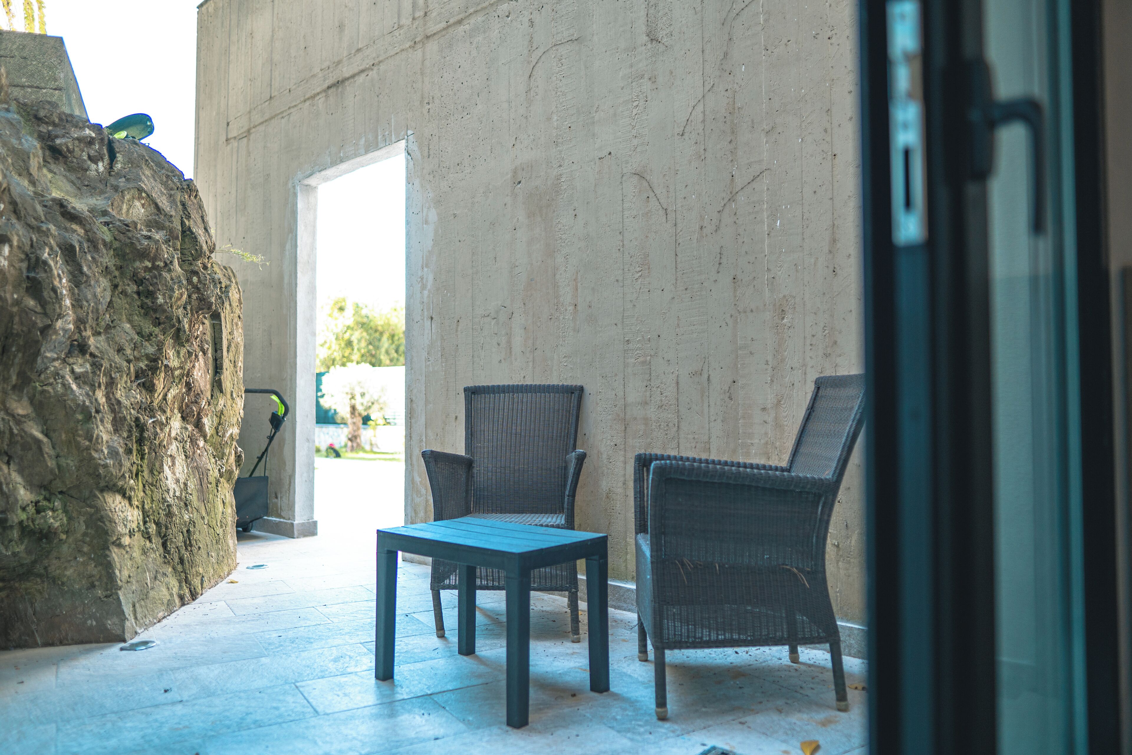 A patio area with two wicker chairs and a table, a large rock, and a concrete wall.