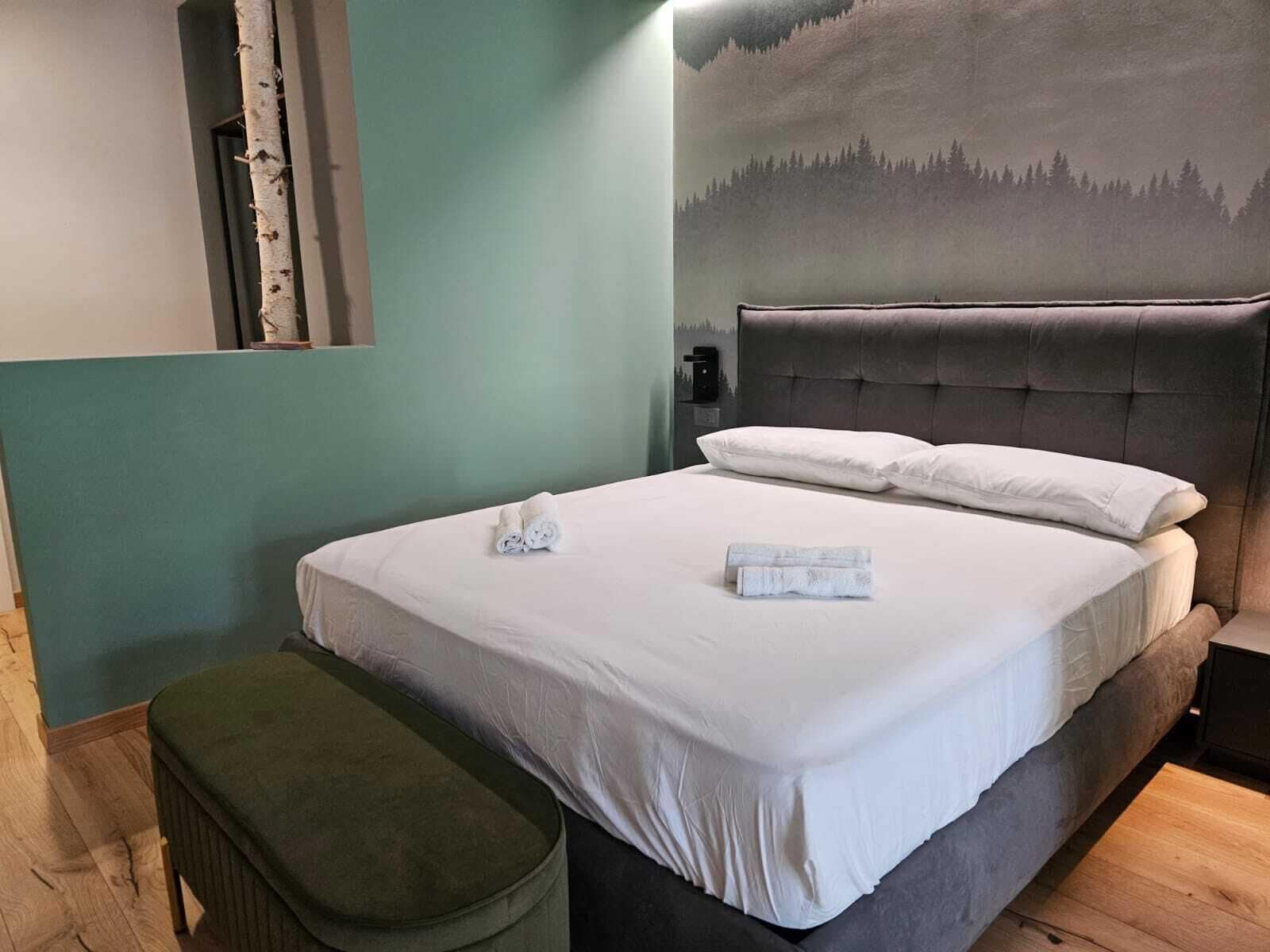 A neatly made bed with white linens and two rolled towels, a green upholstered bench, and a wall-mounted nightstand with a lamp.