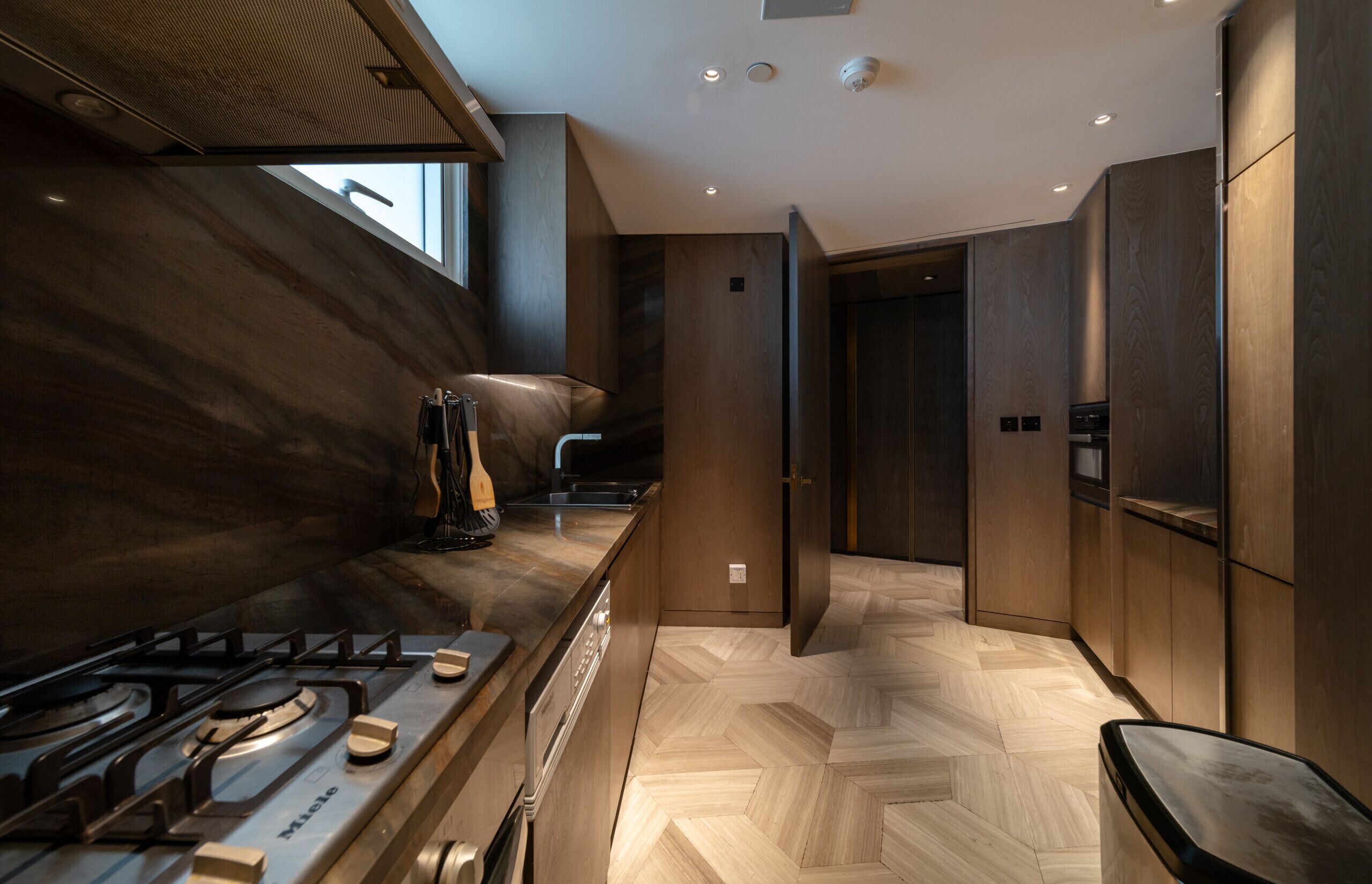 Une cuisine moderne avec un parquet en bois disposé en chevrons, des appareils électroménagers en acier inoxydable et une grande fenêtre qui laisse entrer la lumière naturelle.