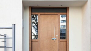 Ein moderner Gebäudeeingang mit einer zweiflügeligen Holztür, einem Metallgitter zur Linken und einer gepflasterten Fläche, die zum Eingang führt.